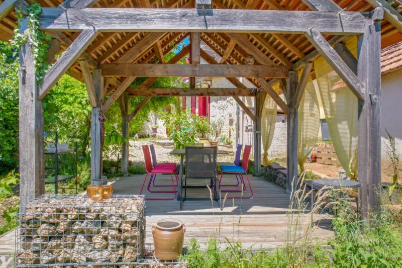 De overdekte pergola met eettafel en gekleurde stoelen, in het groen naast het huis